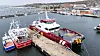 Ett rött och vitt katamaranfartyg ligger förtöjt vid en kaj i en hamn med flera andra båtar i bakgrunden.