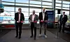 Fyra män står framför en röd invigningsbanderoll i en ljus terminal med stora fönster mot hamnen.