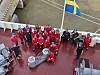 Grupp på fartygsdäck i röda arbetskläder intill vatten och svensk flagga.
