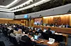 Stor konferenssal med delegater runt bord och en panel längst fram under ett formellt möte.