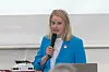 Kvinna i blå jacka talar i mikrofon vid presentation, med modellfartyg och skärm i bakgrunden.