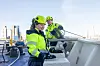 Två personer i gula varselkläder och hjälmar arbetar på ett tak med stad och himmel bakom.