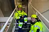 Två arbetare i gula hjälmar sitter på en trappa i en industriell byggnad.