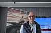 Leende man står framför stora skärmar med bilder av stad och kust.