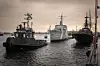 Tre örlogsfartyg förtöjda i en hamn vid vattenytan under en molnig himmel.