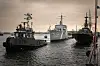 Tre grå marinfartyg ligger för ankar i en hamn med mörkt vatten och molnig himmel.