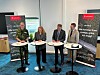 Fyra personer står vid mässbord med broschyrer framför rollups i en konferenslokal.