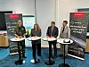 Fyra personer står vid mässbord med broschyrer framför rollups i en konferenslokal.