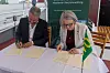 Två personer skriver under papper vid ett bord med en liten grön flagga.