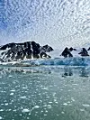 Glaciär och svarta, snötäckta berg vid lugnt vatten under himlen fylld av tunna moln.