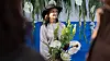 Kvinna i hatt håller ett blomsterarrangemang framför en blå bakgrund och gröna växter.